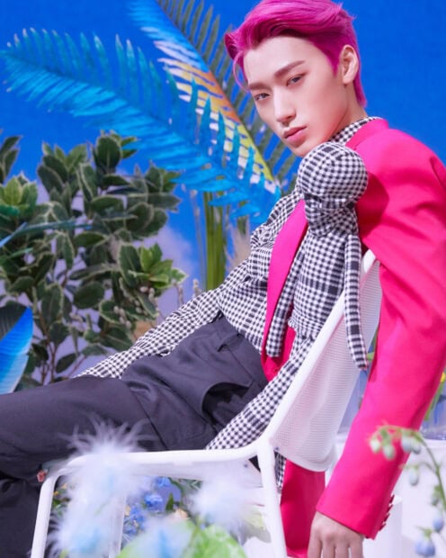 Ateez member with pink hair wearing a checkered outfit reclining on a white chair surrounded by plants