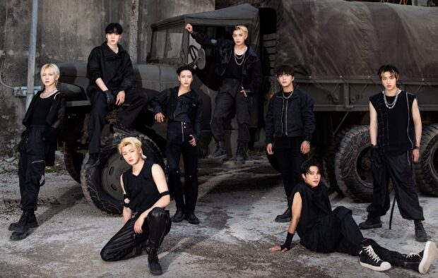 Ateez members posing confidently in black outfits with a rugged military truck in the background