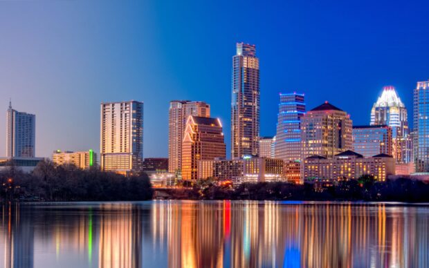 The Austin skyline at dusk with city buildings reflecting on the calm water and clear blue sky