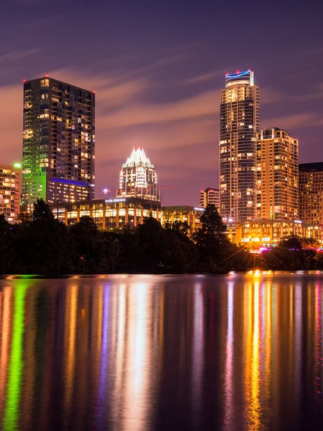 The Austin skyline at night reflecting colorful lights on the water with city buildings towering above trees