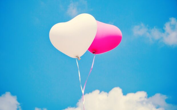 Two heart shaped balloons in white and pink floating in a clear blue sky