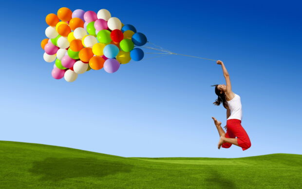A woman jumping joyfully while holding colorful balloons on a clear blue sky above green grass