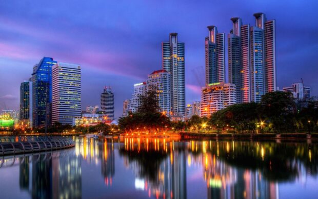Evening cityscape of Bangkok skyline with tall buildings reflecting on calm water at dusk