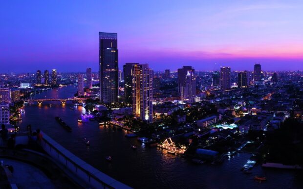 The Bangkok skyline features tall buildings and a river at twilight with vibrant city lights and a purple sky