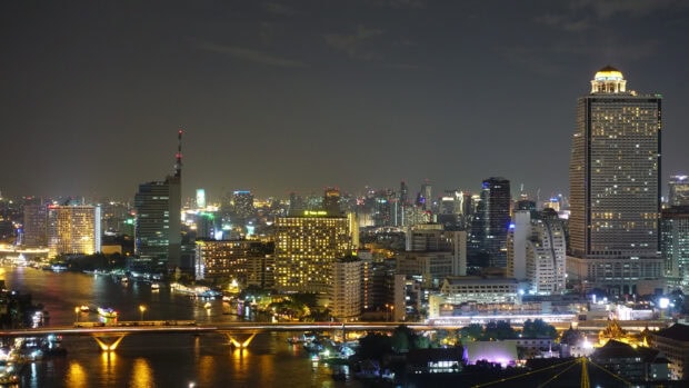 A vibrant Bangkok skyline at night with illuminated buildings and a river reflecting city lights