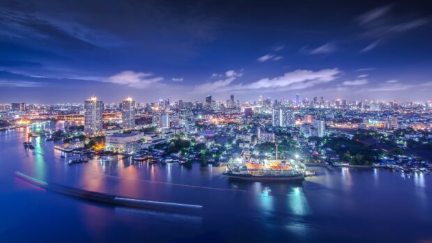 Nighttime cityscape of Bangkok with river and illuminated buildings under a cloudy sky