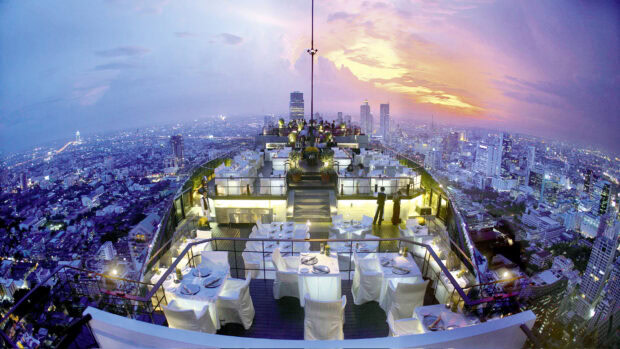 Rooftop dining area in Bangkok cityscape during sunset with vibrant sky and urban buildings