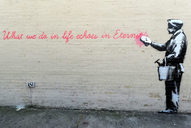 A Banksy artwork showing a painter man with a bucket painting a pink heart on a brick wall