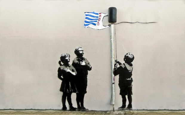 Children standing with hand on chest looking at a flag painted in Banksy style