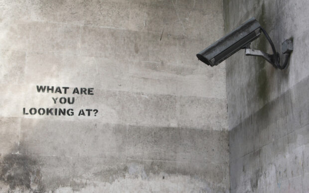 Surveillance camera on wall with Banksy graffiti asking what are you looking at sign