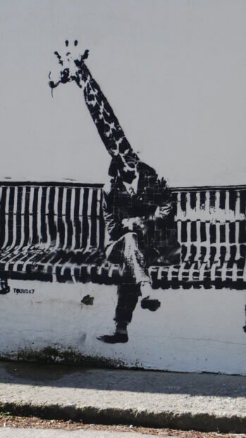 A Banksy artwork showing a man with a giraffe head smoking a pipe sitting on a bench