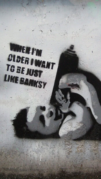 A graffiti stencil art of a child holding a spray can with Banksy text on a rough wall
