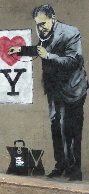 A man with a stethoscope examines a heart symbol on a wall in Banksy graffiti style