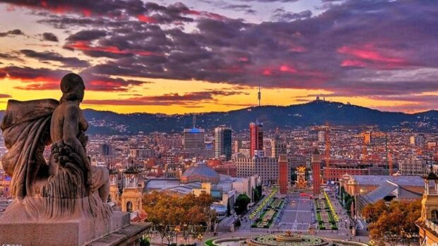 Cityscape of Barcelona with a statue in the foreground under a colorful sunset sky