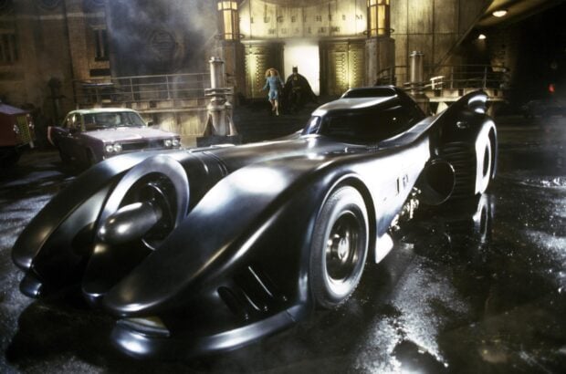 A sleek Batmobile parked in front of an industrial building with Batman and a woman in the background