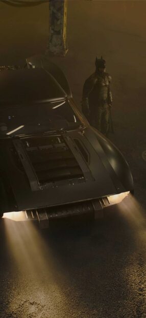 Batman standing near a dark Batmobile with headlights on in a dimly lit area