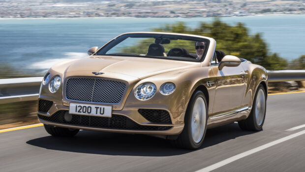 A golden Bentley Continental convertible driving on a coastal road with a scenic ocean view