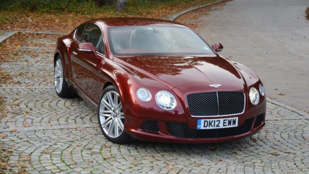 A red Bentley Continental car parked on a cobblestone driveway surrounded by fallen leaves