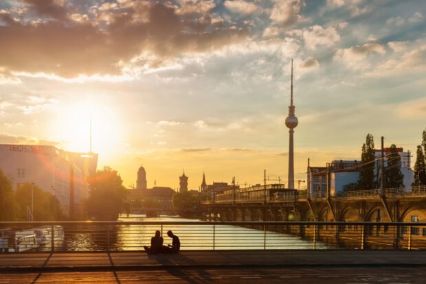Sunset view of Berlin skyline featuring the television tower and river with two people sitting on a bridge