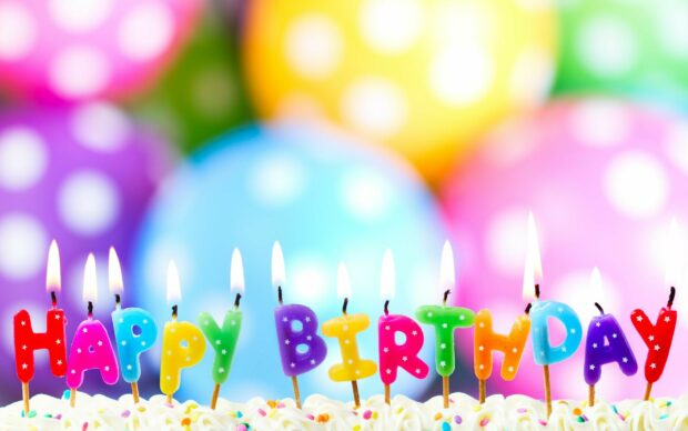 Colorful happy birthday candles lit on top of a cake with blurred balloons background