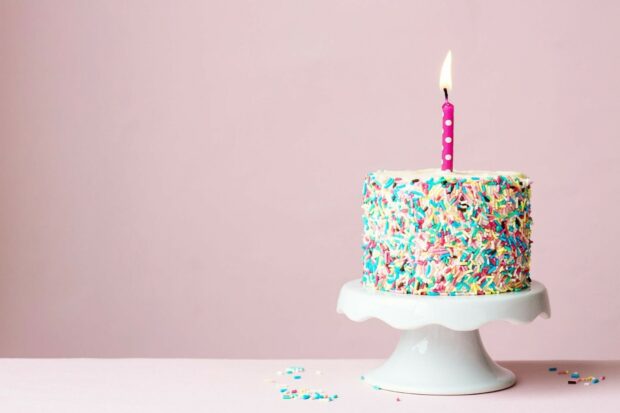 A birthday cake with colorful sprinkles and a lit candle on a white cake stand