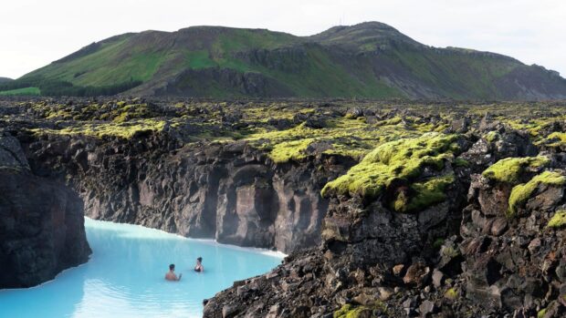 A serene volcanic landscape with mossy rocks and two people bathing in blue lagoon Iceland
