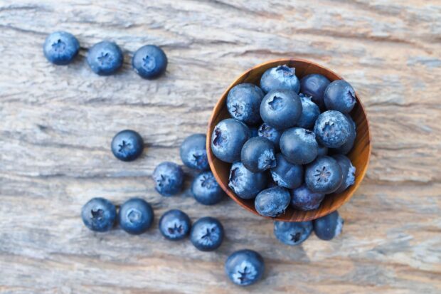 A wooden bowl filled with fresh blueberry on a rustic wooden surface with scattered blueberry around