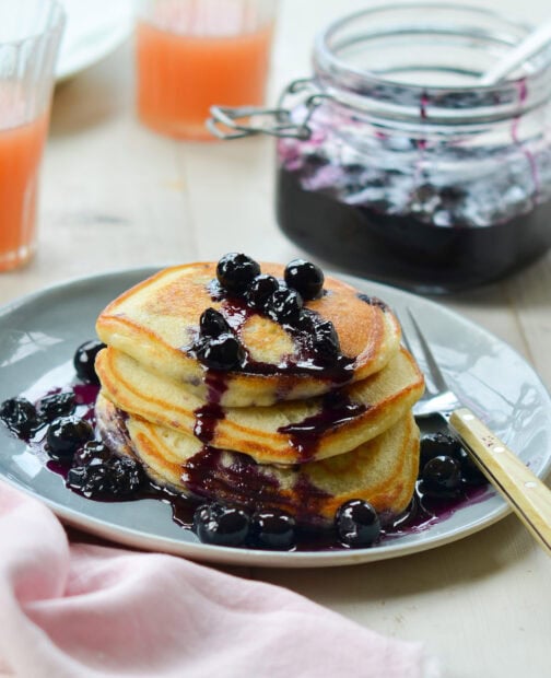 A stack of pancakes topped with blueberry compote and fresh blueberry around on a plate