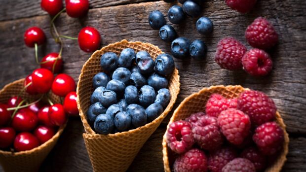 A close up of blueberry in a waffle cone on a wooden surface with cherries and raspberries nearby