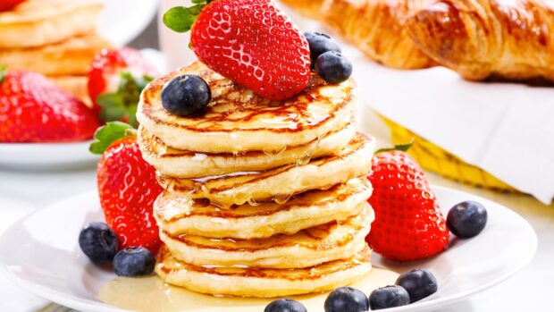 A stack of pancakes topped with fresh blueberry and strawberry surrounded by more berries on a white plate