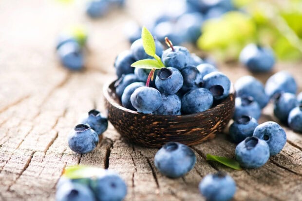 Fresh blueberry fruit in a wooden bowl on a rustic wooden surface with green leaves