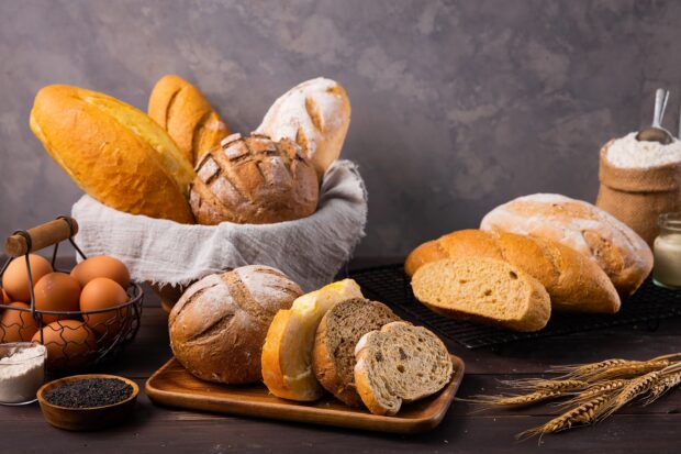 Various fresh bread loaves arranged on a wooden table with wheat and eggs as ingredients