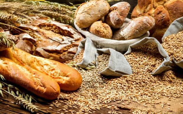 Fresh loaves of bread surrounded by sacks of wheat grains and ear stalks on a wooden surface