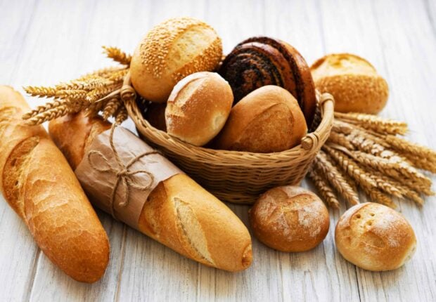 A basket of assorted fresh bread loaves and wheat stalks on a wooden surface