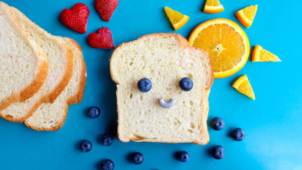 A slice of bread with blueberries as eyes and a pasta smile surrounded by fruit pieces on a blue surface