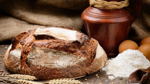 A rustic loaf of bread with wheat flour eggs and a clay jug on a wooden table