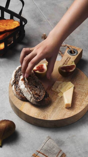 A hand picking a slice of whole grain bread on a wooden board with figs and cheese slices
