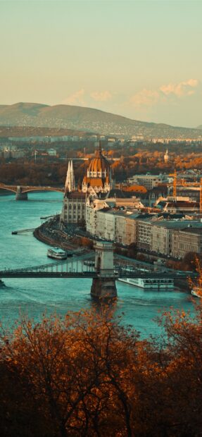 The historic Parliament building in Budapest is viewed from above with autumn trees and the Danube River
