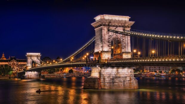 Historic Budapest Bridge illuminated at night over calm river with cityscape in the background