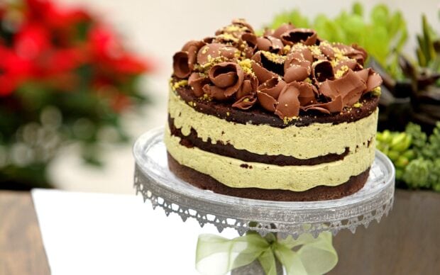 A layered cake with green frosting and chocolate curls on top displayed on a glass cake stand
