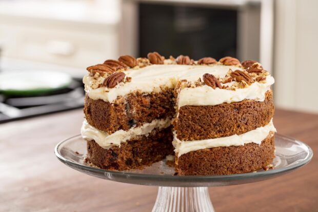 A close up of a carrot cake with cream cheese frosting and pecans on a glass cake stand