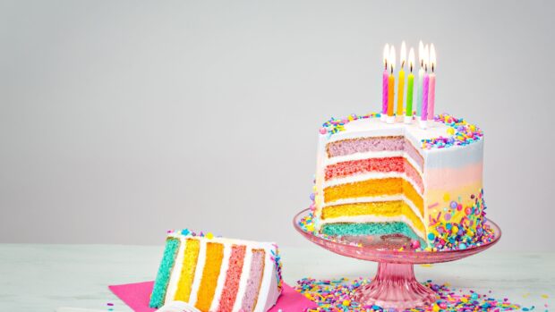A colorful rainbow cake with candles and sprinkles on a pink cake stand