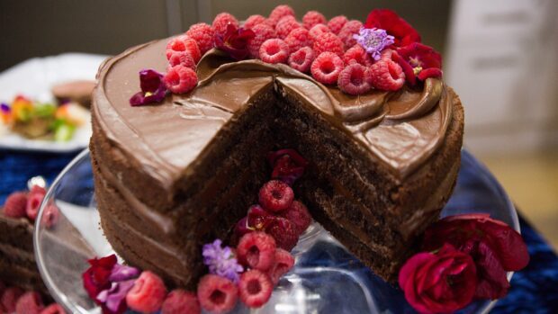A chocolate cake decorated with raspberries and flowers on a glass stand
