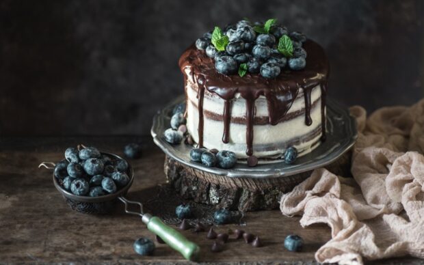 A delicious cake topped with fresh blueberries and chocolate drizzle on a rustic wooden surface