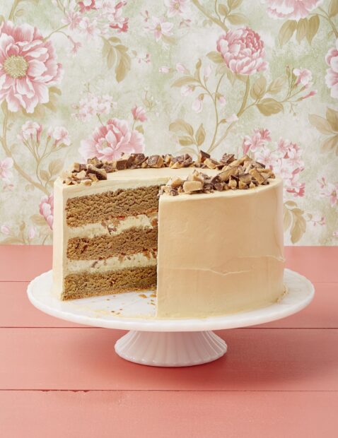A layered cake with peanut butter frosting and chopped candy pieces on top displayed on a white cake stand