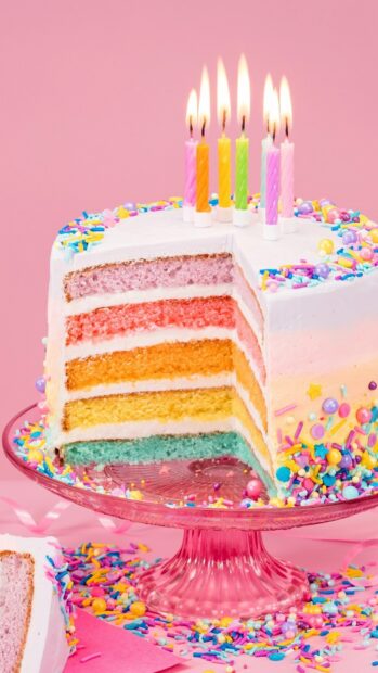 A colorful cake with layered sponge and sprinkles on a glass stand with lit candles