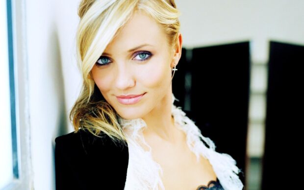 Cameron Diaz with blonde hair and blue eyes wearing a black jacket and white lace top leaning against a wall
