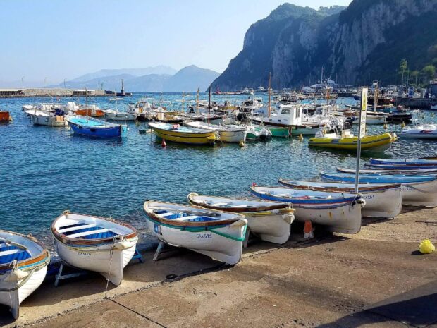 Small boats lined up along the shore at Capri Island harbor with clear blue sea and rocky cliffs in the background