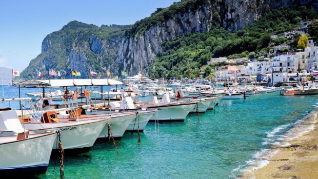 A scenic view of Capri island boats docked in clear turquoise water near rocky cliffs and hillside buildings