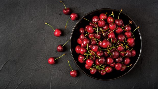 Fresh cherry fruits arranged in a black bowl with some cherries scattered on a dark textured surface